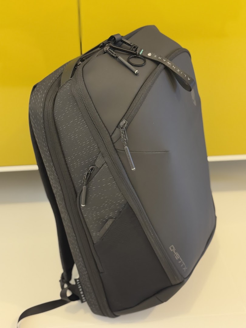 A sleek black Alienware backpack with multiple compartments and branding, displayed against a yellow wall.