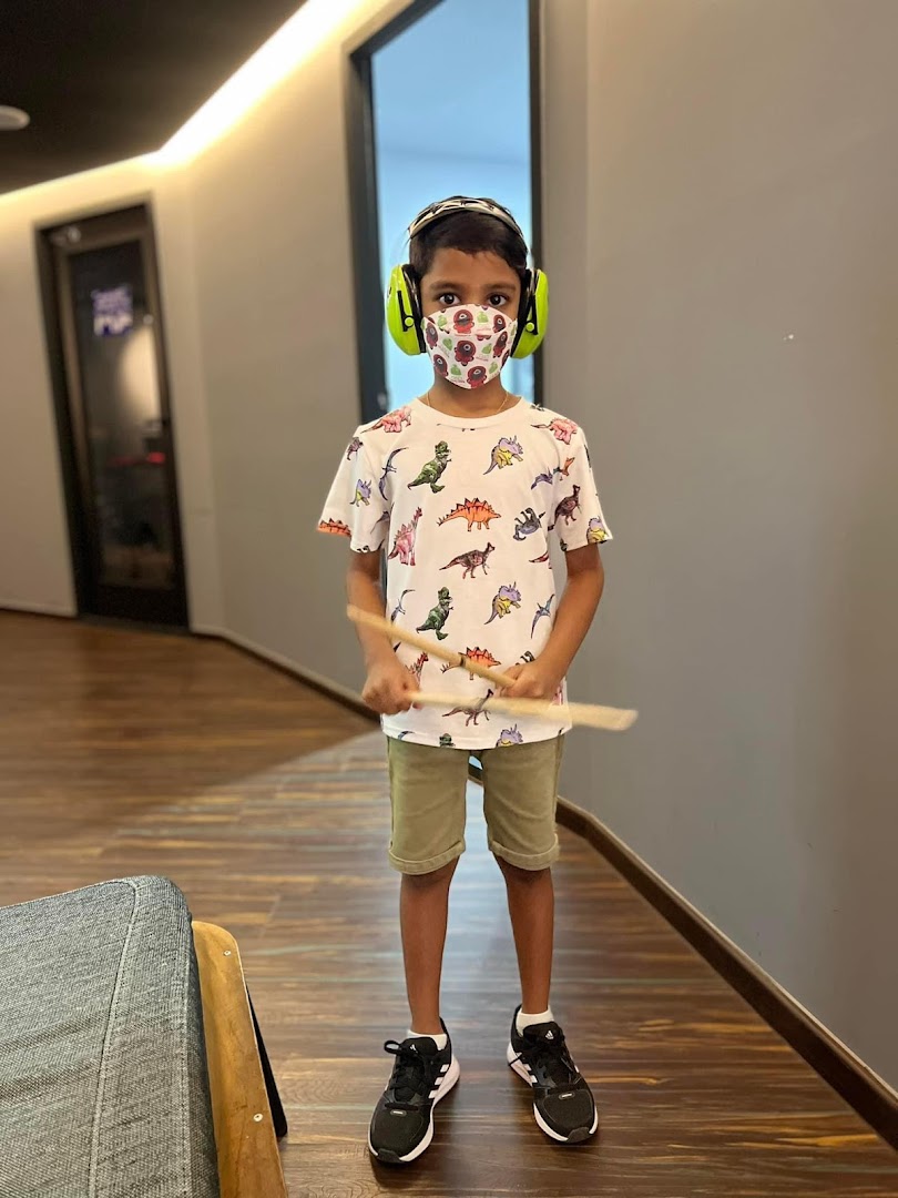 A young boy wearing ear protection and a mask, holding drumsticks in a studio hallway.
