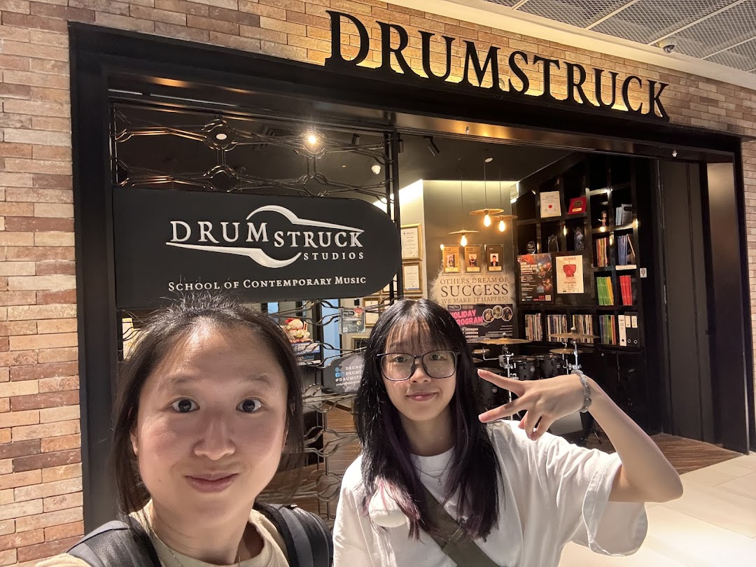 Two individuals pose in front of Drumstruck Studios, a school of contemporary music.