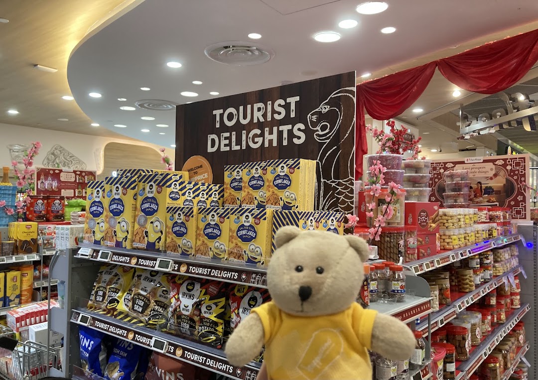 A supermarket aisle featuring 'Tourist Delights' products, including snacks and local specialties, with a teddy bear in the foreground.