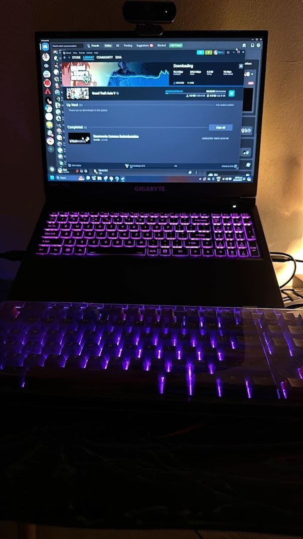 A gaming laptop and external keyboard with vibrant purple backlighting, displaying a game download on Steam.