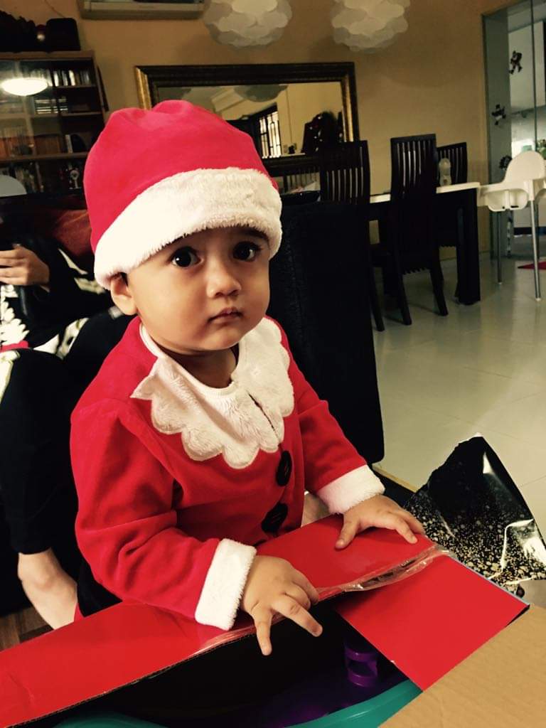 A cute baby in a Santa costume opening a red box, looking at the camera.