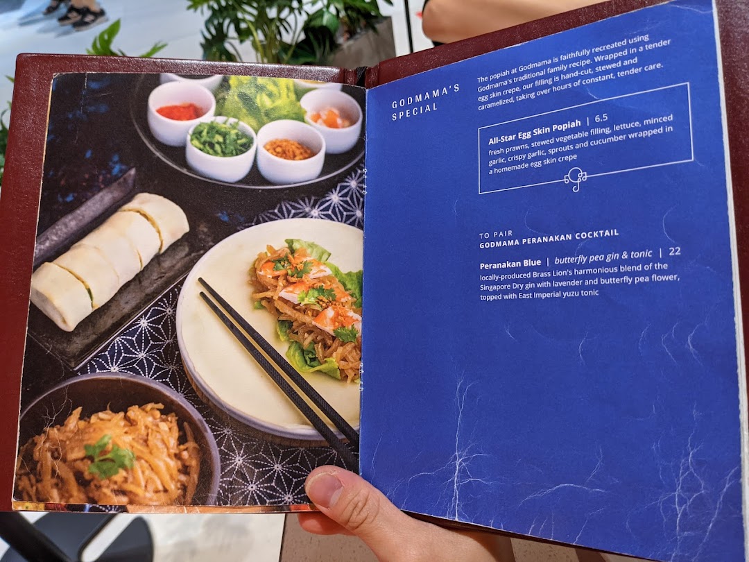 An open menu from Godmama restaurant, showcasing a popiah dish and a Peranakan cocktail.