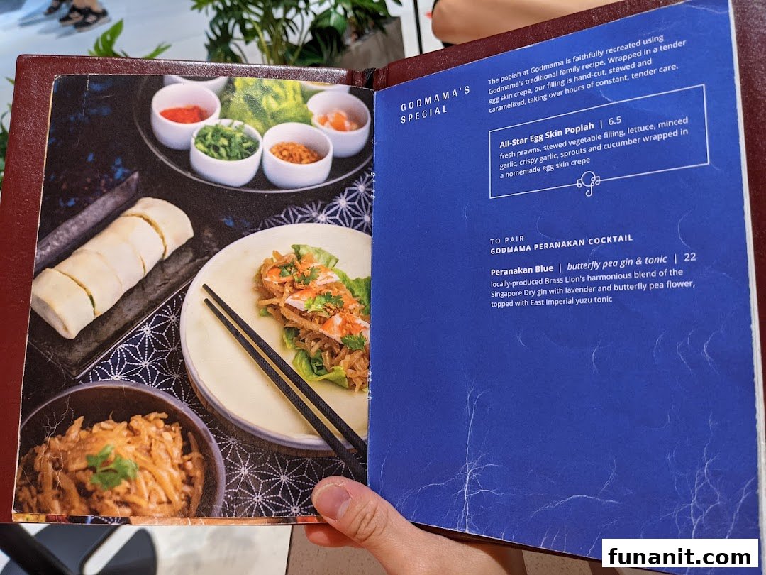 An open menu from Godmama restaurant, showcasing a popiah dish and a Peranakan cocktail.