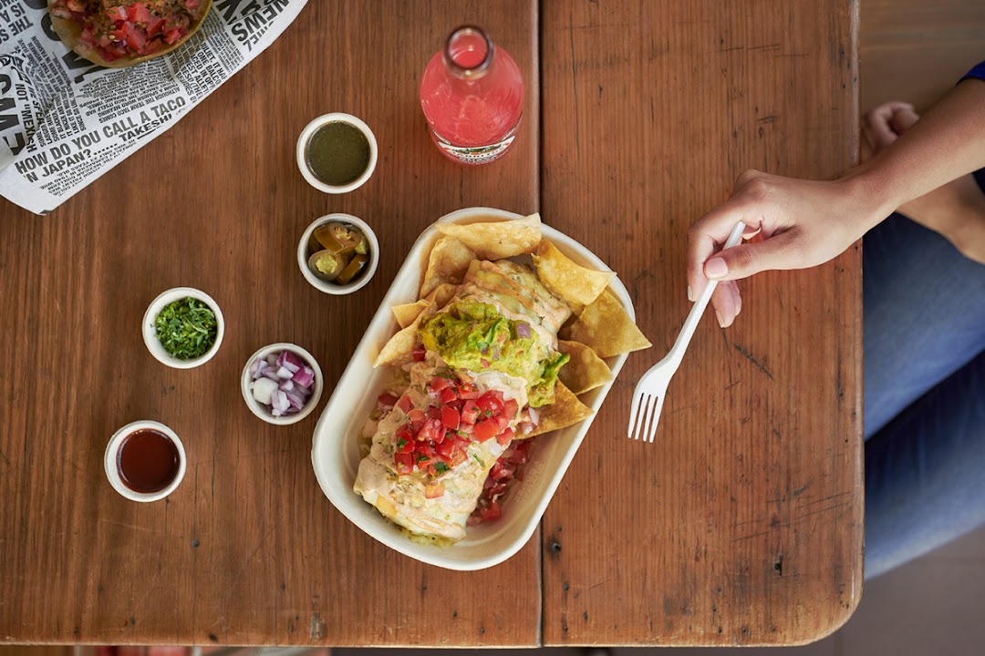 Overhead view of a person about to enjoy a large burrito bowl with chips and condiments on a wooden table.