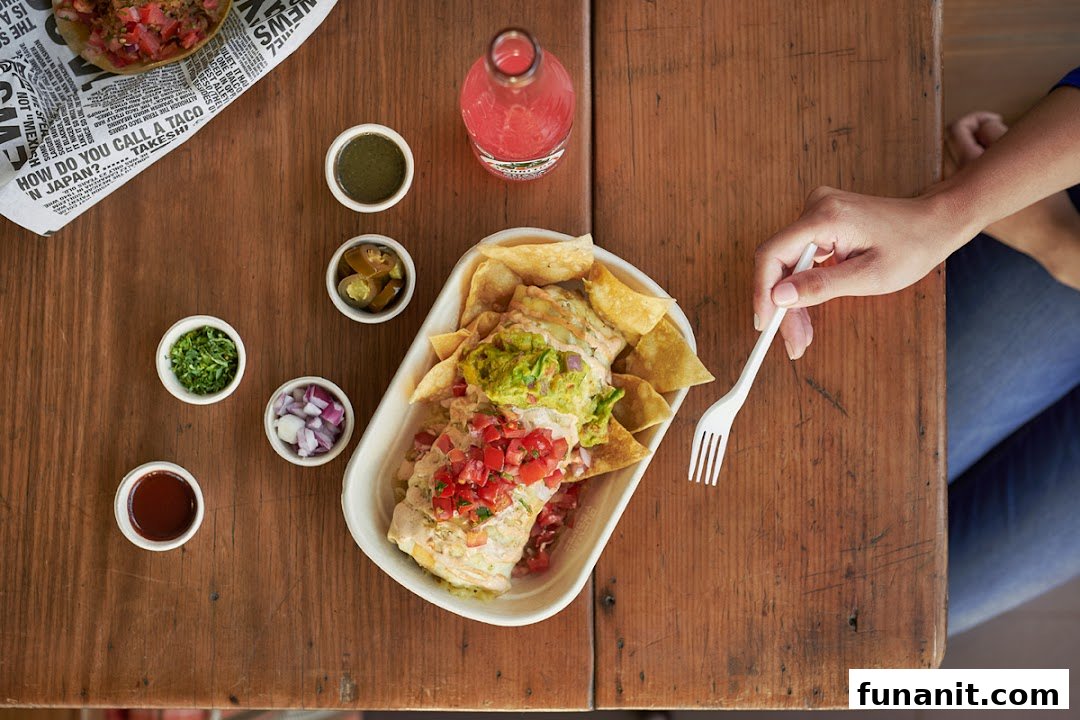 Overhead view of a person about to enjoy a large burrito bowl with chips and condiments on a wooden table.