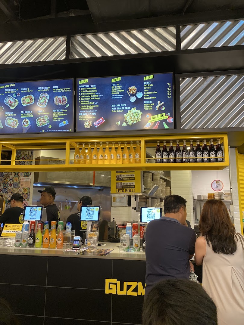 Customers at Guzman y Gomez counter with digital menu boards displaying various Mexican food and drink options.