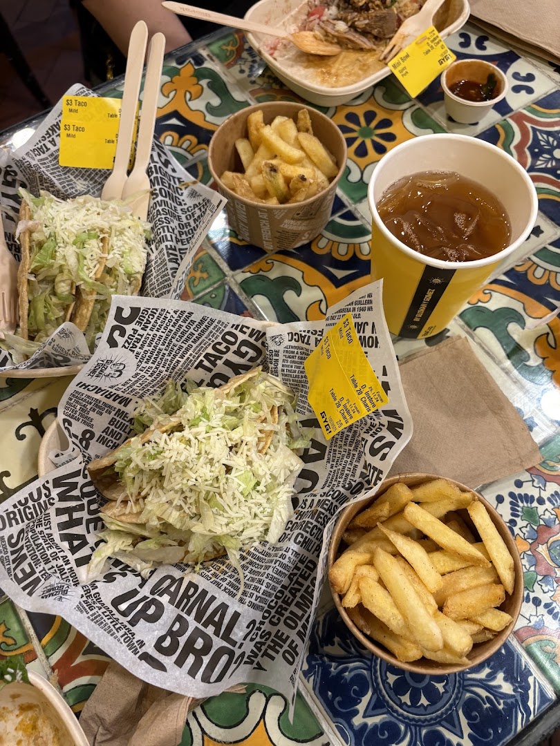 A vibrant close-up of delicious tacos, fries, and a drink from Guzman y Gomez on a colorful tiled table.