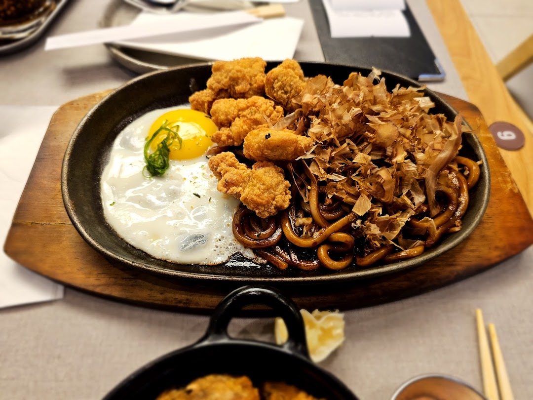 A sizzling hot plate featuring Japanese noodles, crispy fried chicken, a sunny-side-up egg, and bonito flakes.