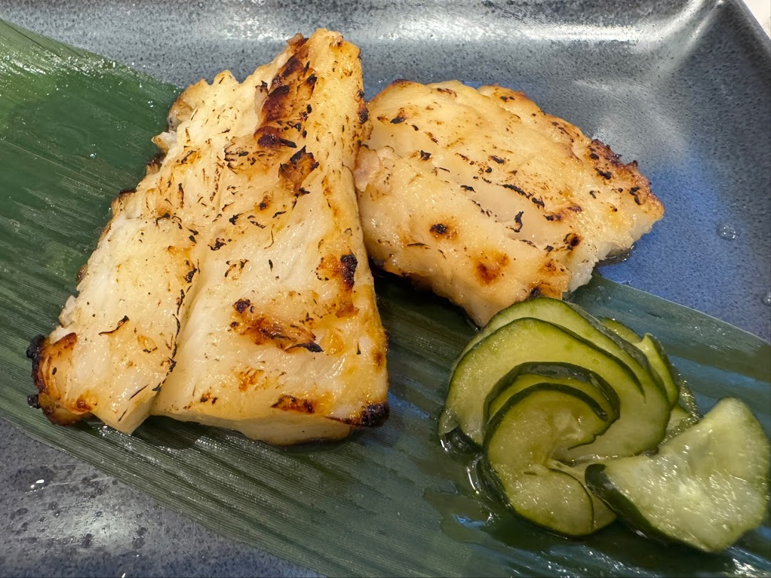 Two pieces of perfectly grilled fish served on a banana leaf with sliced pickled cucumbers.