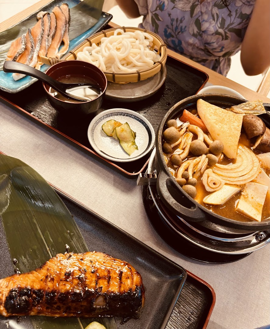 A table laden with various Japanese dishes, including grilled salmon, sukiyaki hotpot, udon, and pickled cucumbers.