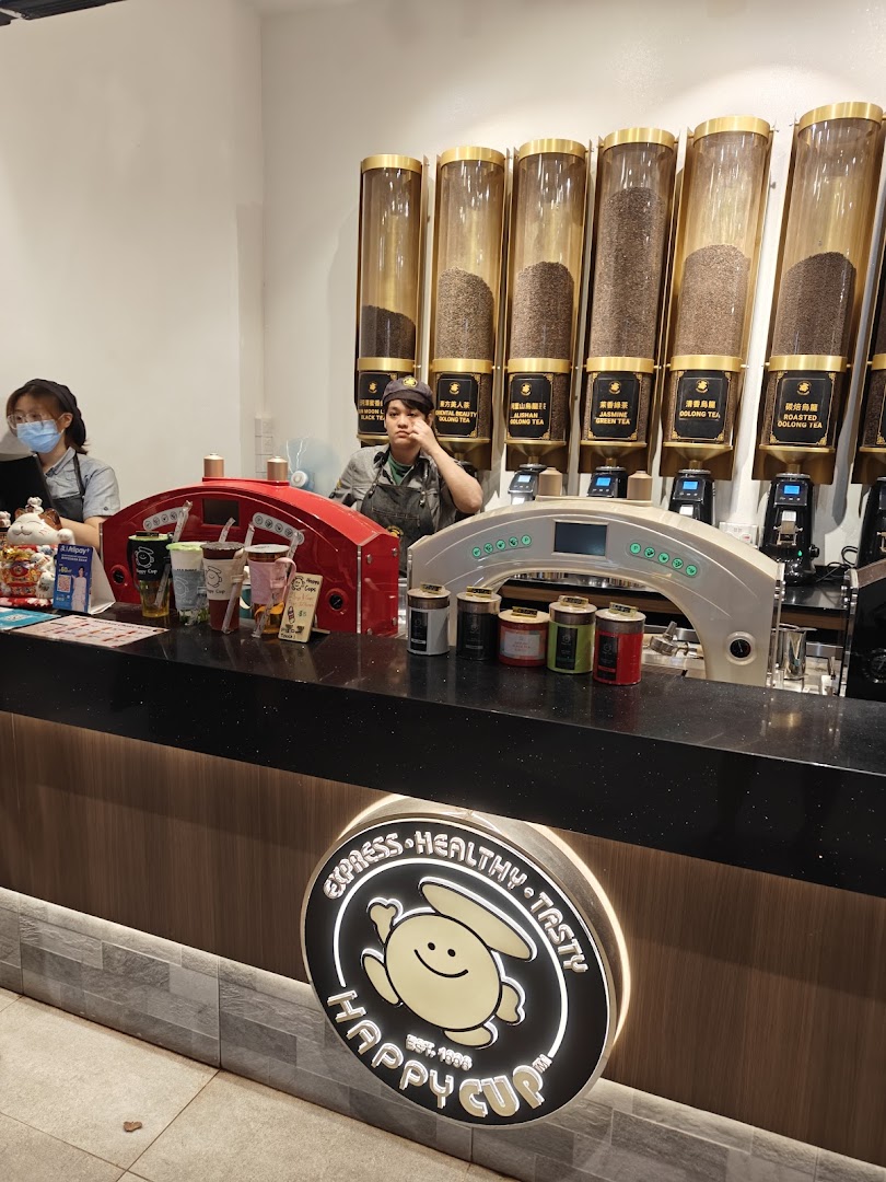 Two staff members at the counter of Happy Cup, with large tea dispensers and brewing equipment visible.