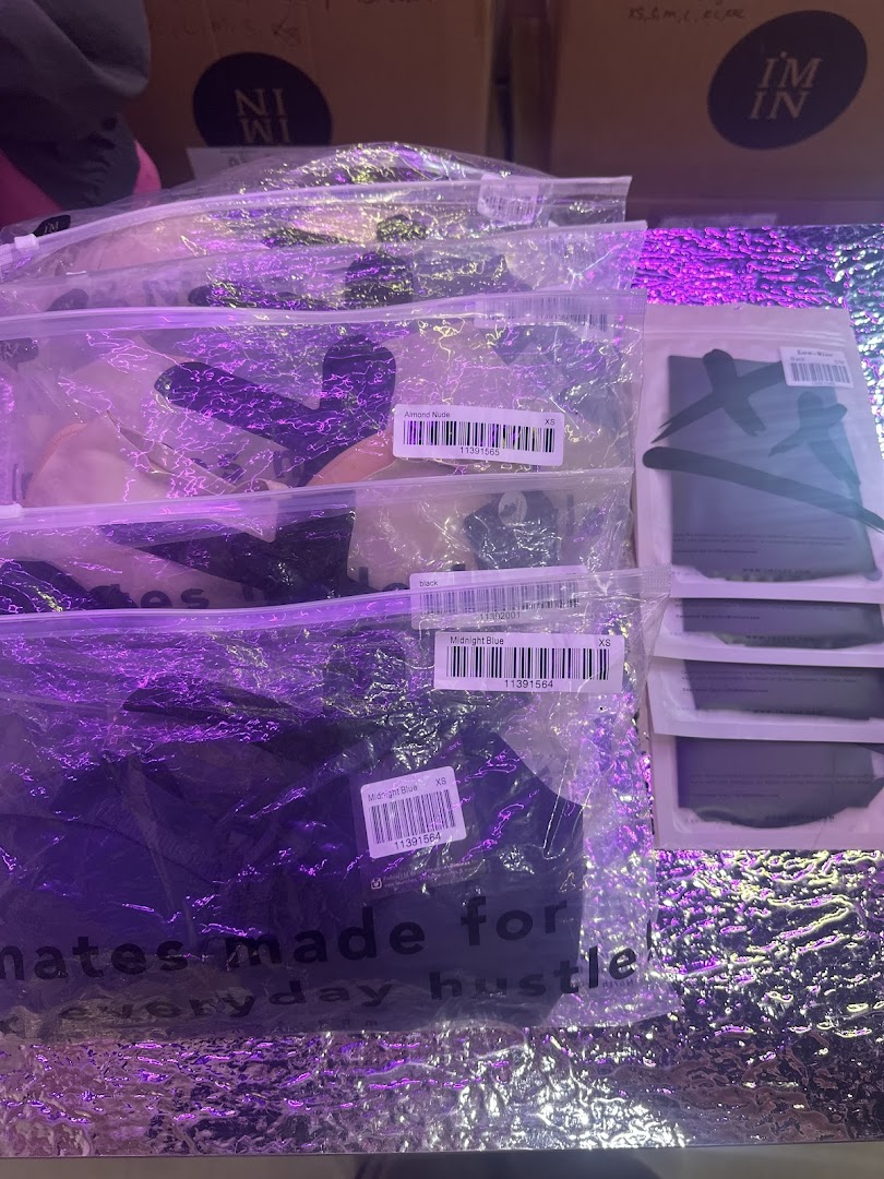 Packaged apparel items in various colors, including 'Almond Nude' and 'Midnight Blue', displayed under purple lighting.