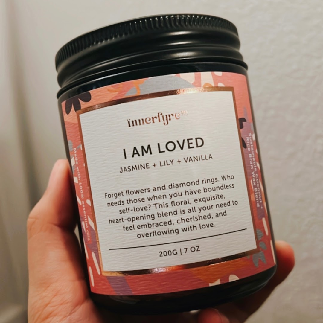 A hand holds an Innerfyre Co. 'I Am Loved' candle jar with jasmine, lily, and vanilla scent.