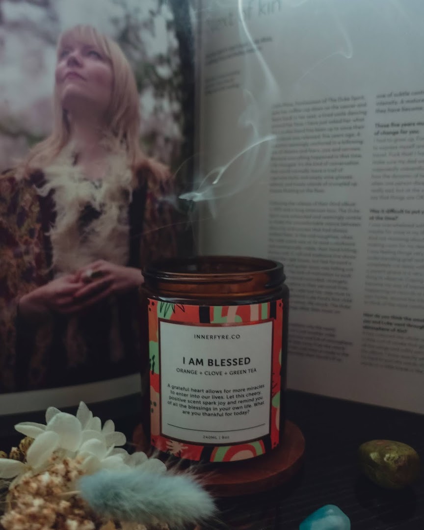 A lit Innerfyre Co 'I Am Blessed' candle with smoke, featuring orange, clove, and green tea scents, alongside a book and decor.