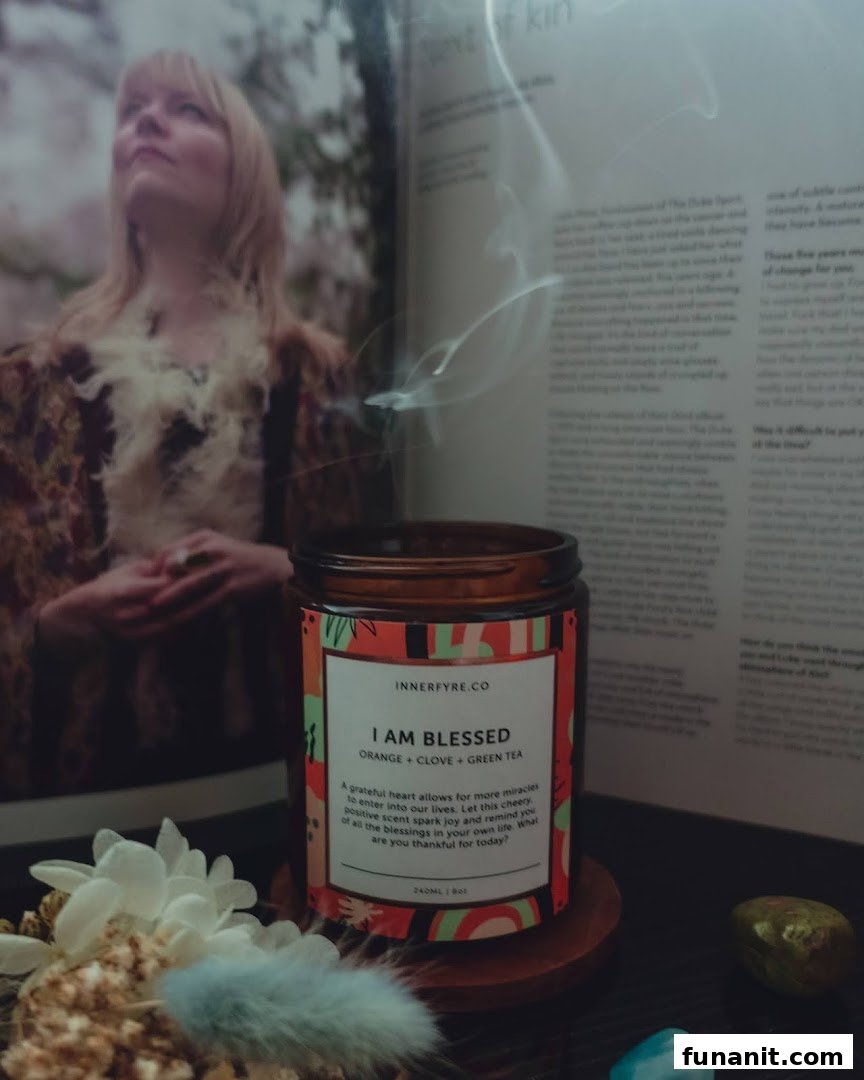 A lit Innerfyre Co 'I Am Blessed' candle with smoke, featuring orange, clove, and green tea scents, alongside a book and decor.