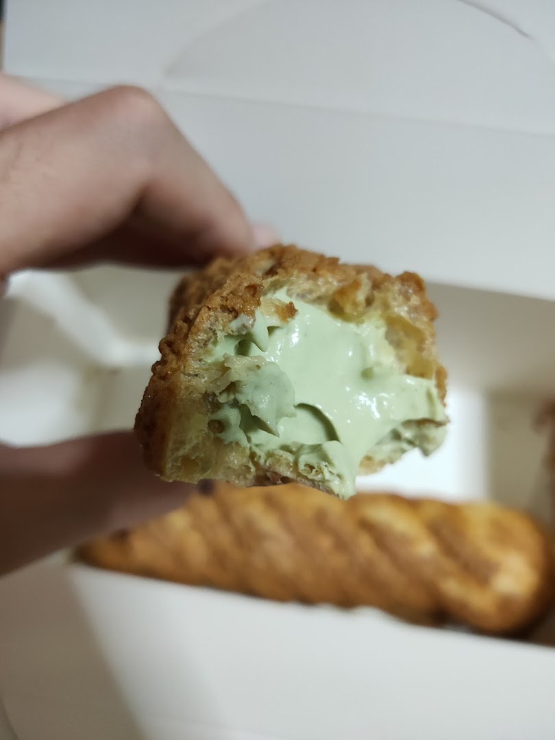 A hand holds a choux pastry with vibrant green filling, showcasing its creamy texture inside a box.