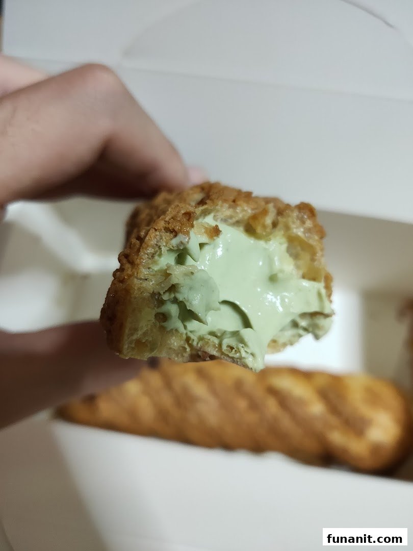 A hand holds a choux pastry with vibrant green filling, showcasing its creamy texture inside a box.