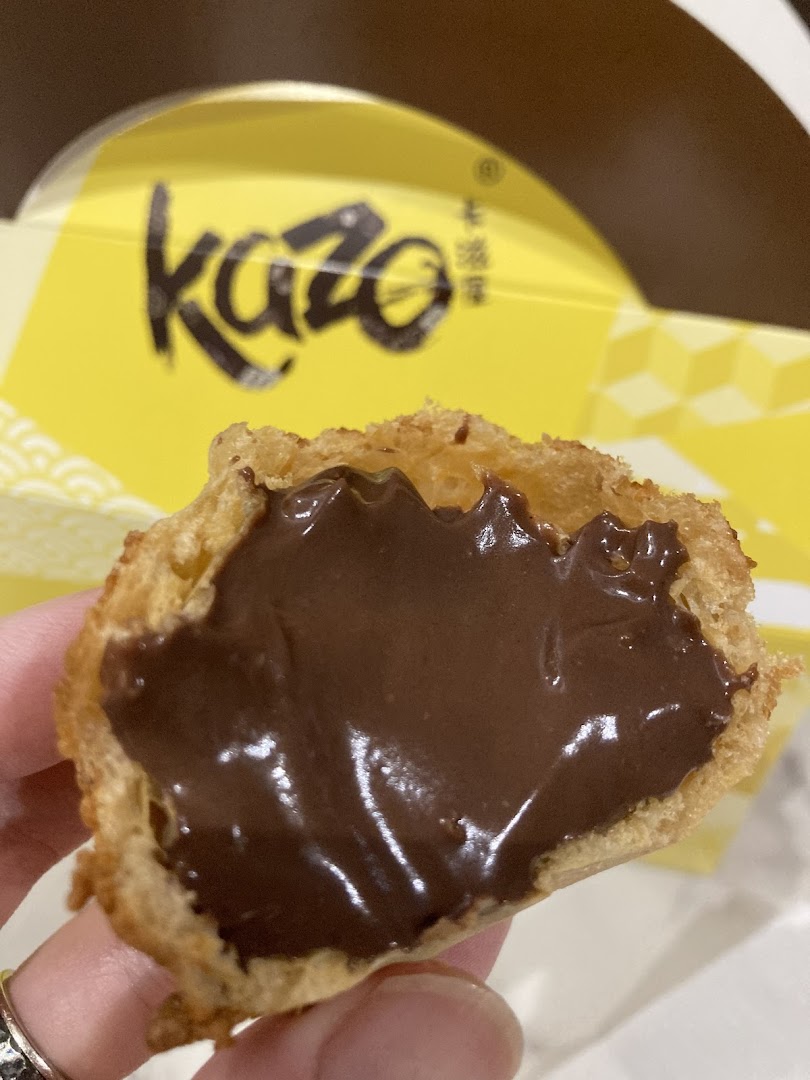 A hand holds a Kazo cream puff, revealing its rich chocolate cream filling, with the brand's yellow box visible.