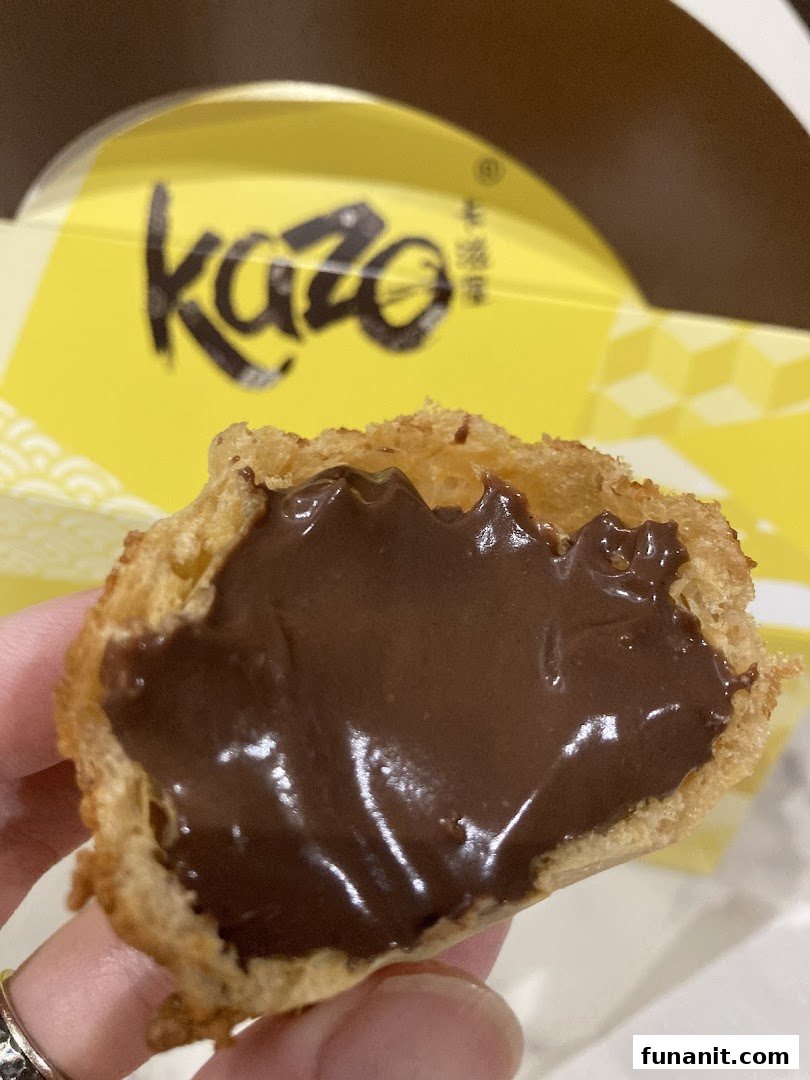 A hand holds a Kazo cream puff, revealing its rich chocolate cream filling, with the brand's yellow box visible.
