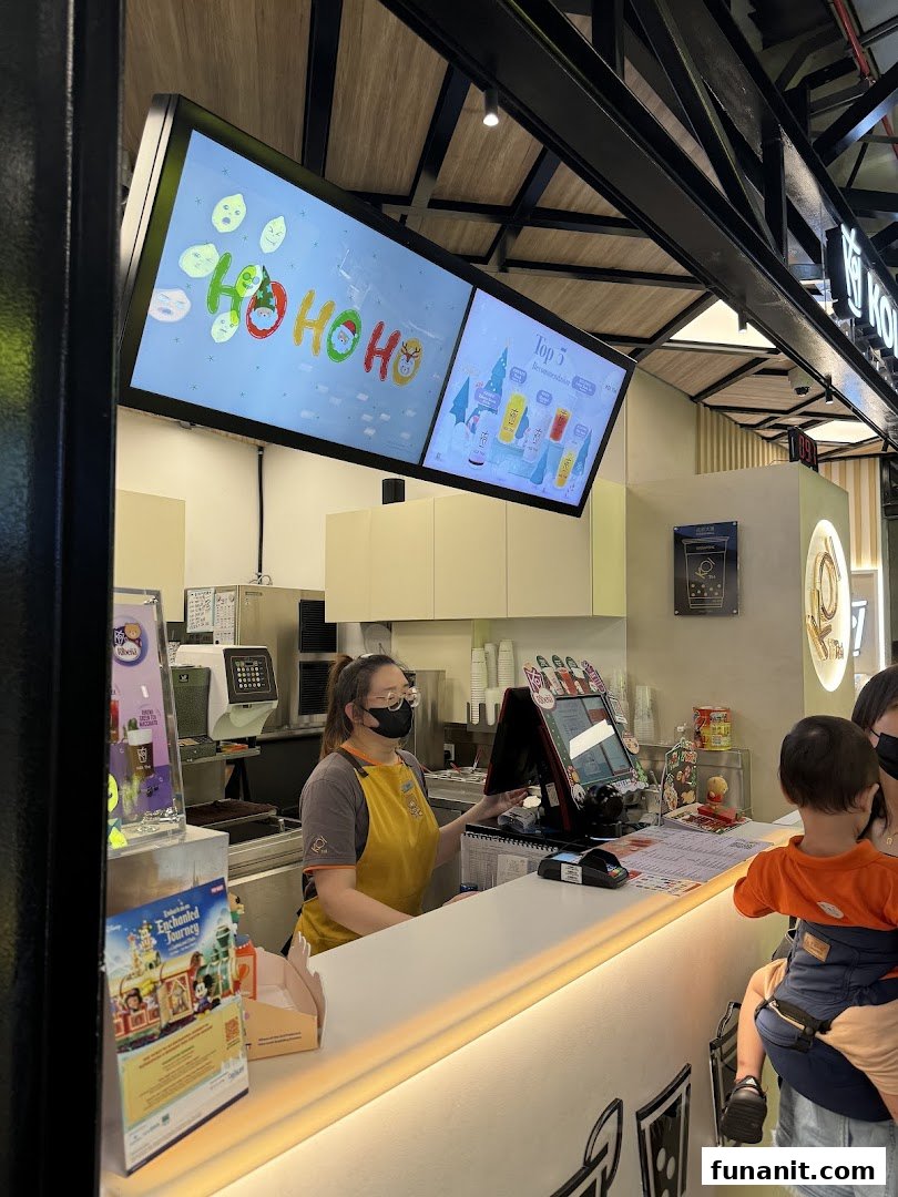A KOI Thé staff member serves a customer at the brightly lit counter, with digital displays above.