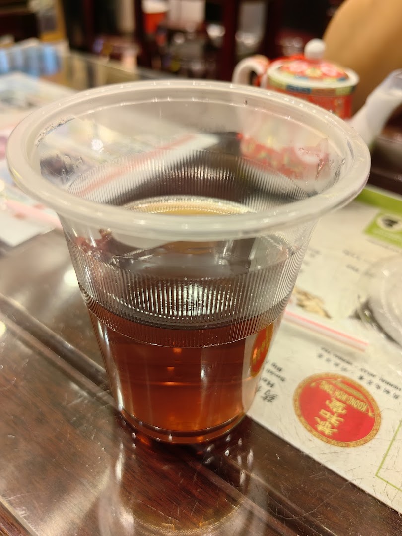A clear plastic cup filled with dark herbal tea on a wooden table at Koong Woh Tong.