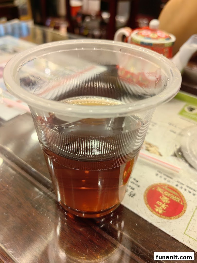 A clear plastic cup filled with dark herbal tea on a wooden table at Koong Woh Tong.