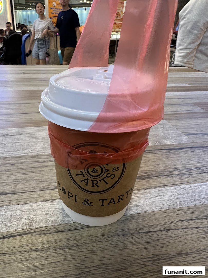 A Kopi & Tarts takeaway cup with a red plastic carrier sits on a wooden table.