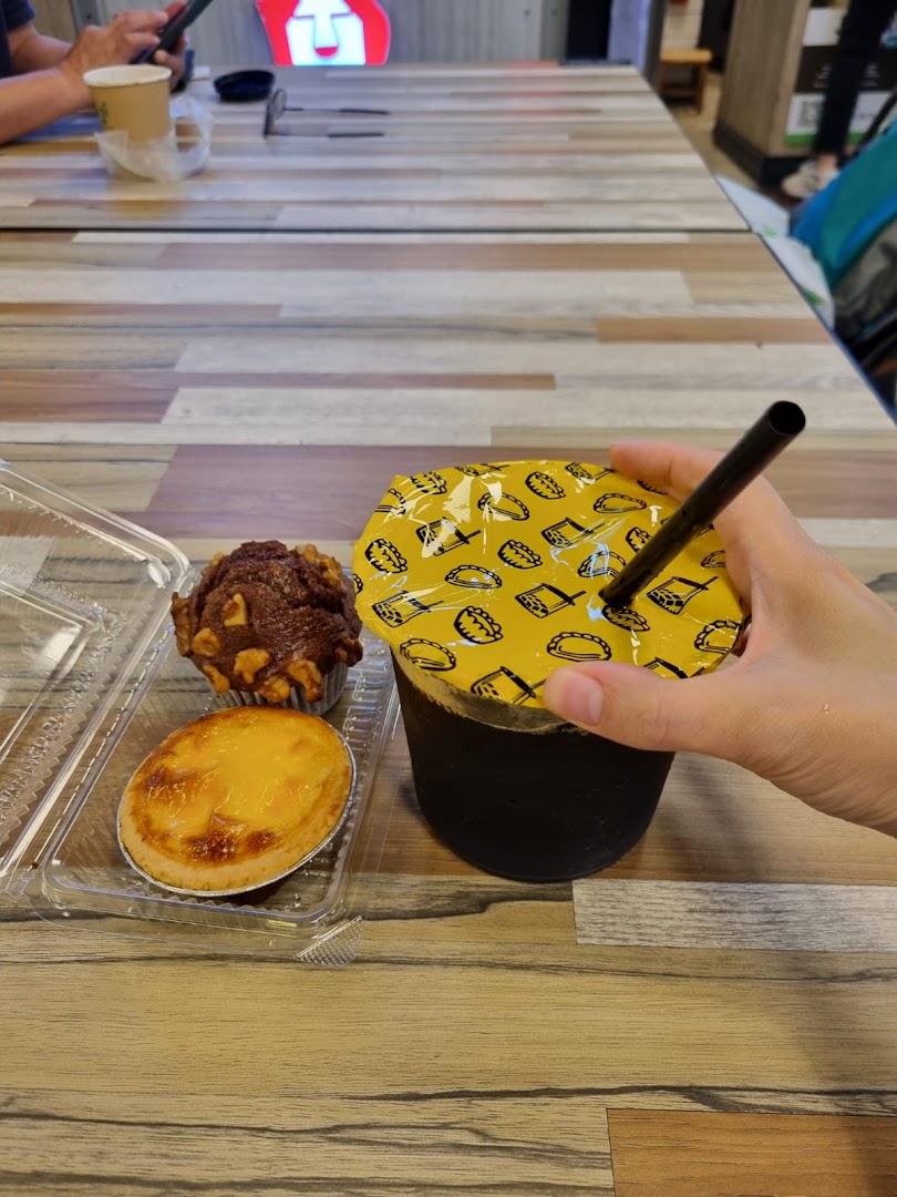A hand holds an iced drink next to an egg tart and muffin on a wooden table.