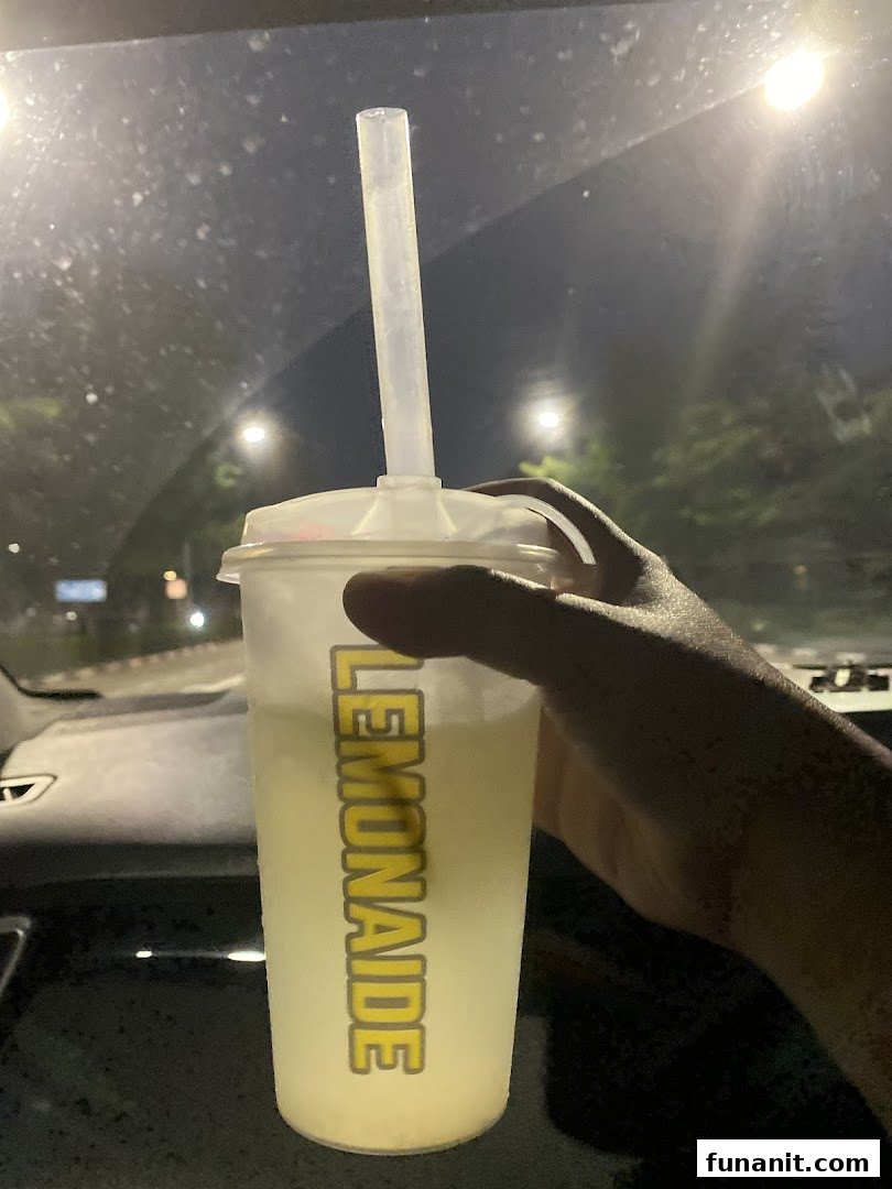 A hand holds a refreshing cup of Lemonaide with a straw, illuminated at night.