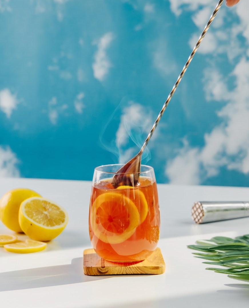 A refreshing glass of hot lemon tea with citrus slices and a stirring spoon, set against a blue sky.