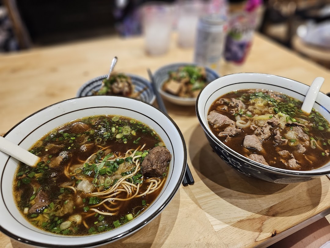 Two steaming bowls of rich beef noodles and side dishes on a wooden table, showcasing a delicious meal.