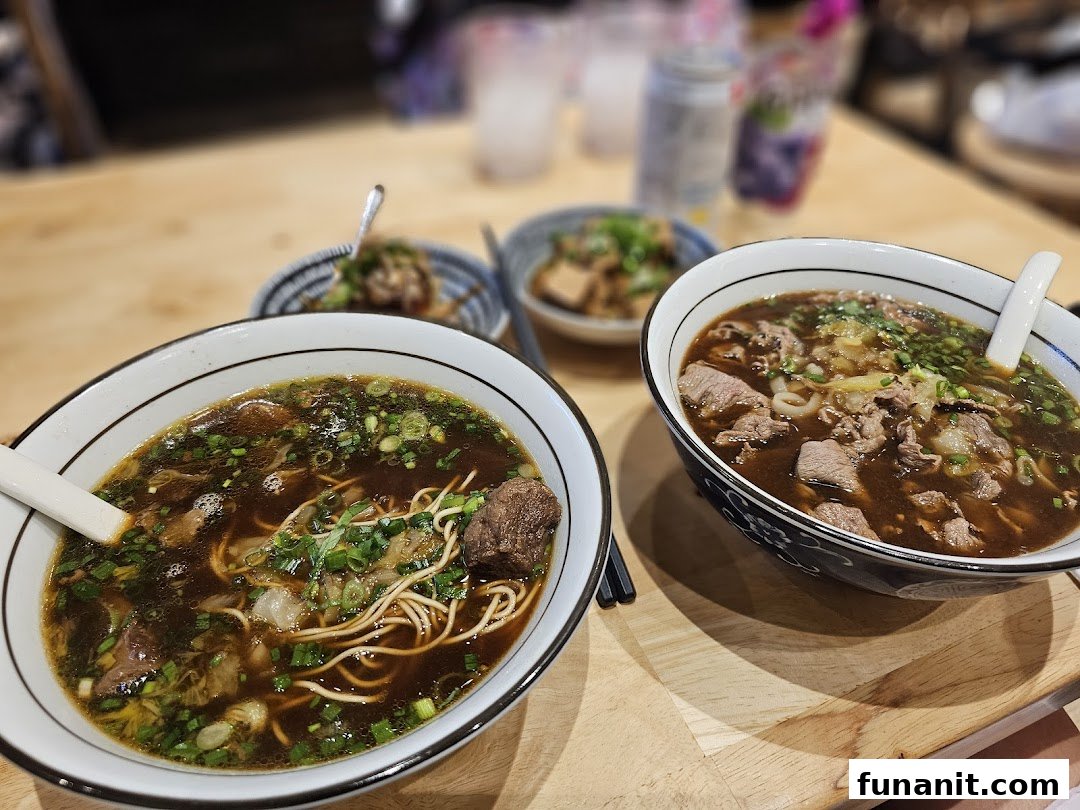 Two steaming bowls of rich beef noodles and side dishes on a wooden table, showcasing a delicious meal.