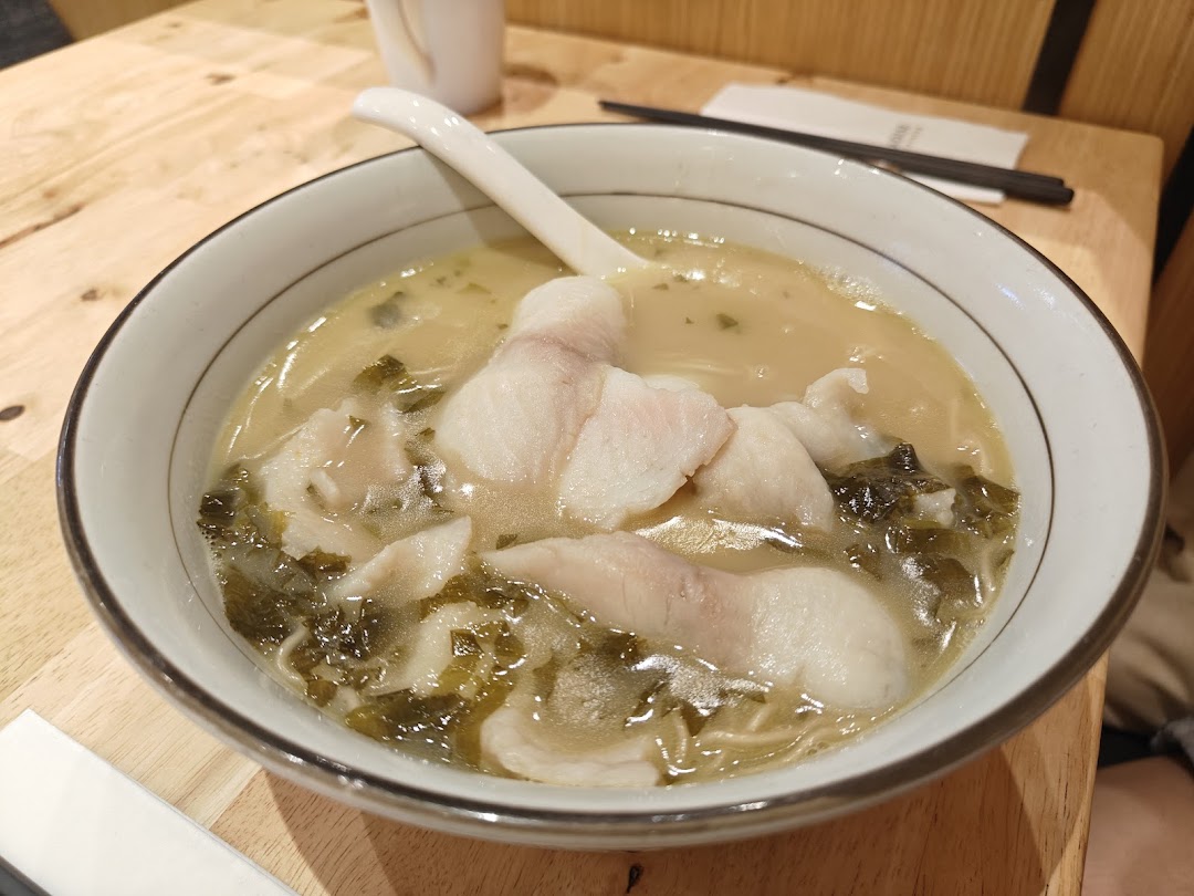 A close-up of a bowl of fish noodle soup with pickled vegetables on a wooden table.