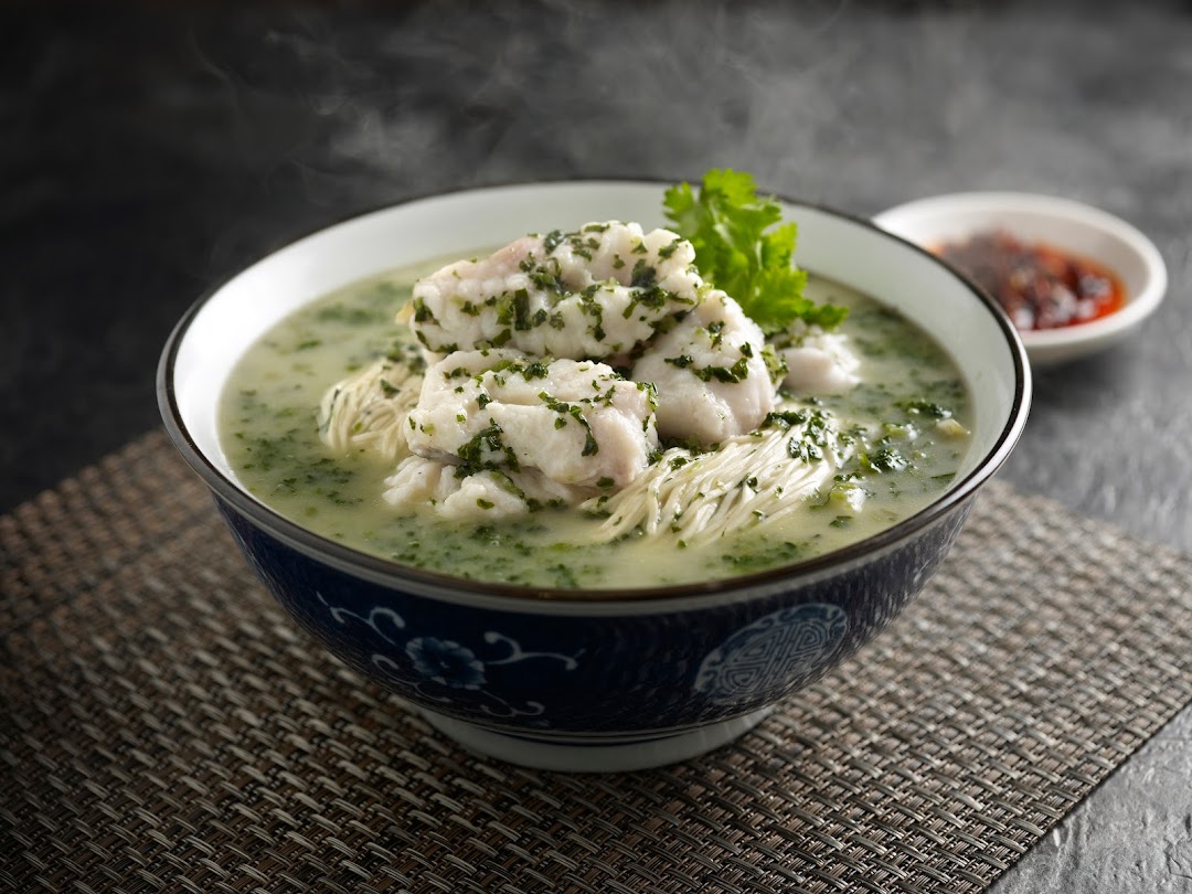 A steaming bowl of fish and noodle soup with green herbs, served with a side of chili.
