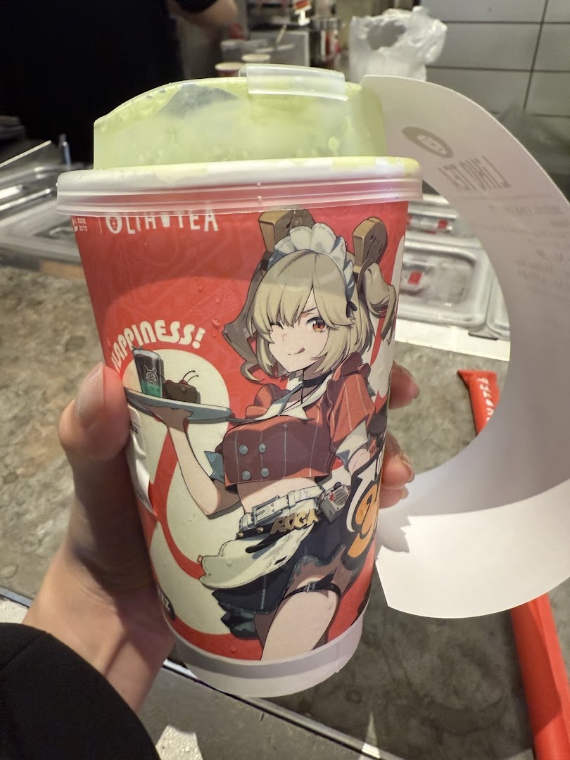 A hand holds a LiHO TEA cup featuring a unique anime character design and a green-colored drink.