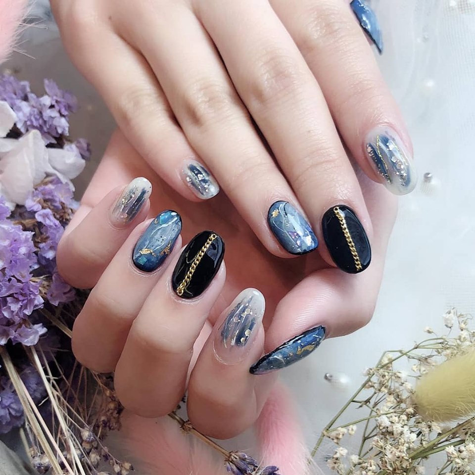 Close-up of hands showcasing intricate dark and light nail art with gold accents and chains.