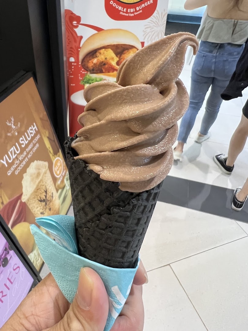 A hand holds a chocolate soft serve in a black waffle cone, with promotional posters in the background.