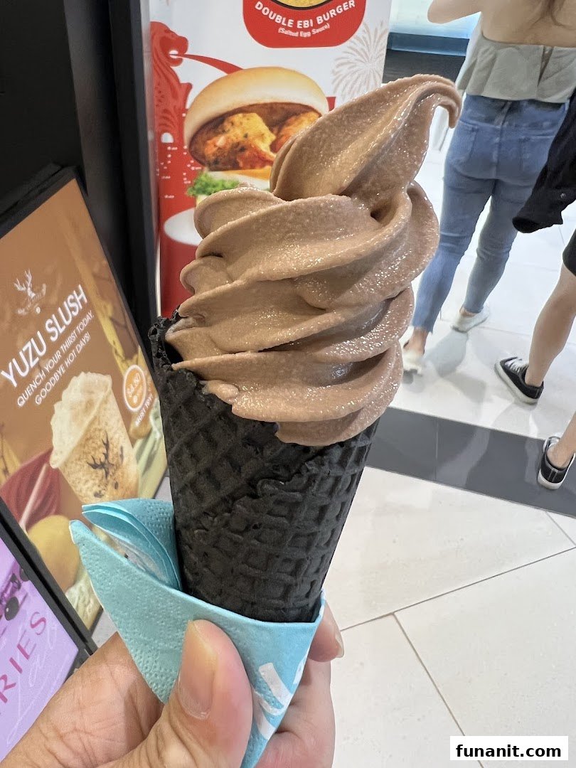 A hand holds a chocolate soft serve in a black waffle cone, with promotional posters in the background.