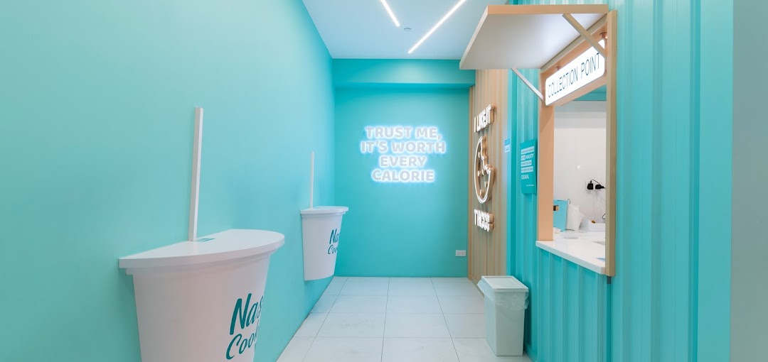 Vibrant turquoise interior of Nasty Cookie with a neon sign and large cup decor leading to a collection point.