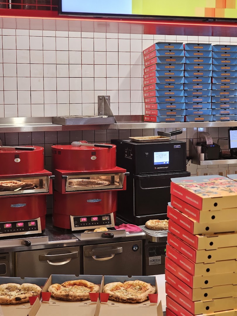 Inside Now Pizza, showing ovens, fresh pizzas, and stacks of branded pizza boxes.