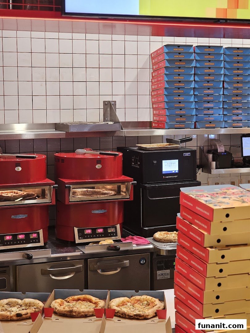 Inside Now Pizza, showing ovens, fresh pizzas, and stacks of branded pizza boxes.