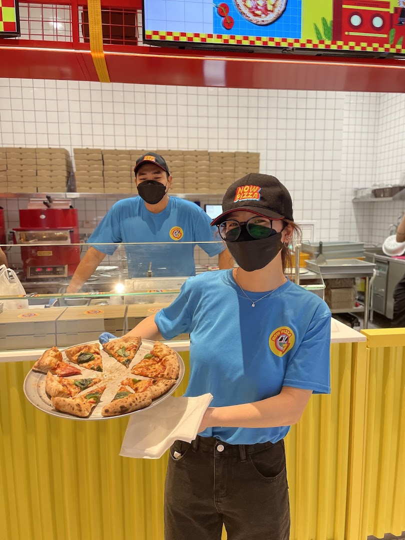 Now Pizza staff member holds a tray of freshly sliced pizza, with another staff in the background.