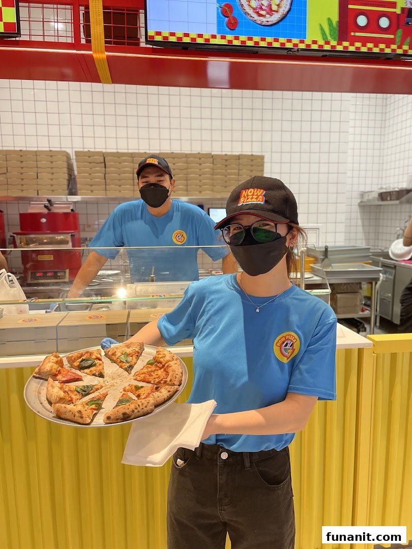 Now Pizza staff member holds a tray of freshly sliced pizza, with another staff in the background.
