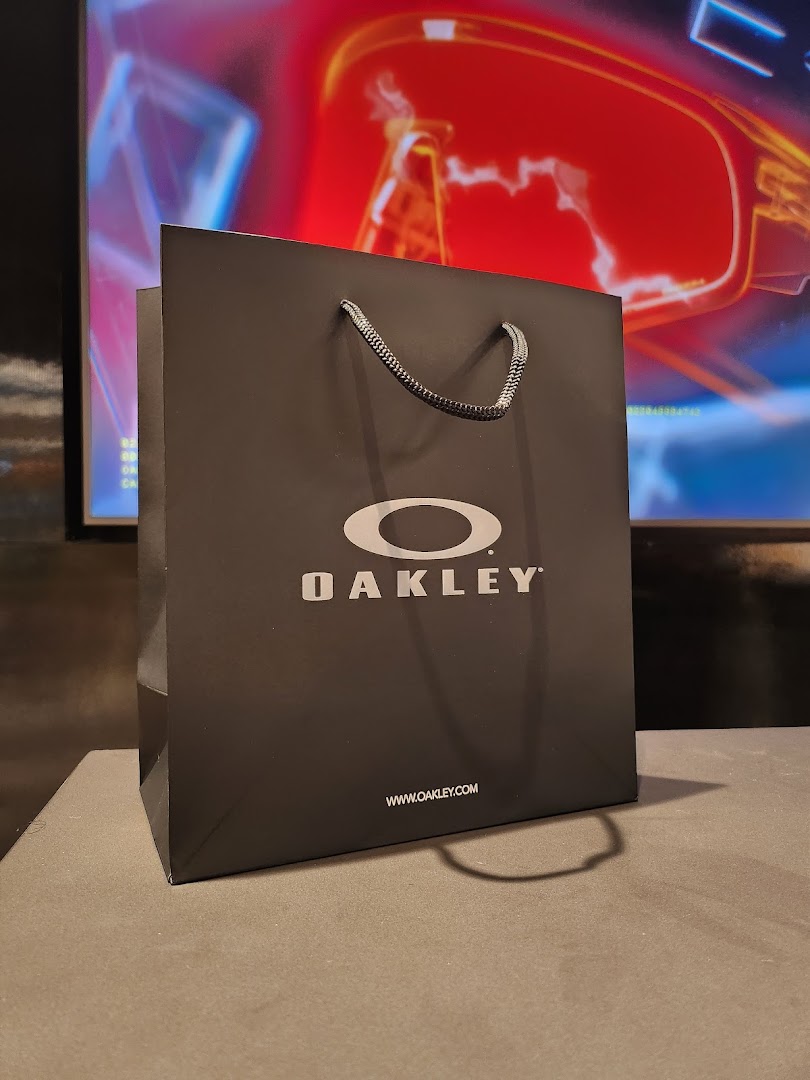 A black Oakley shopping bag with the brand logo and website, set against a blurred background.