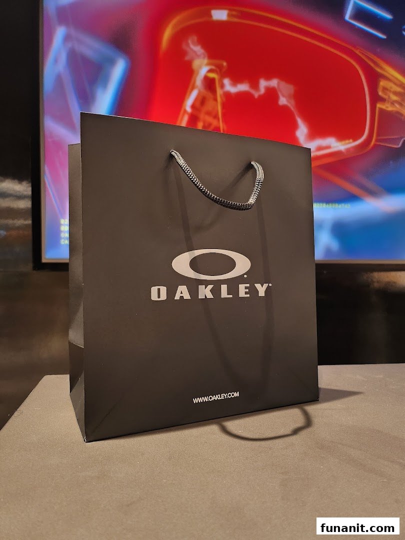 A black Oakley shopping bag with the brand logo and website, set against a blurred background.