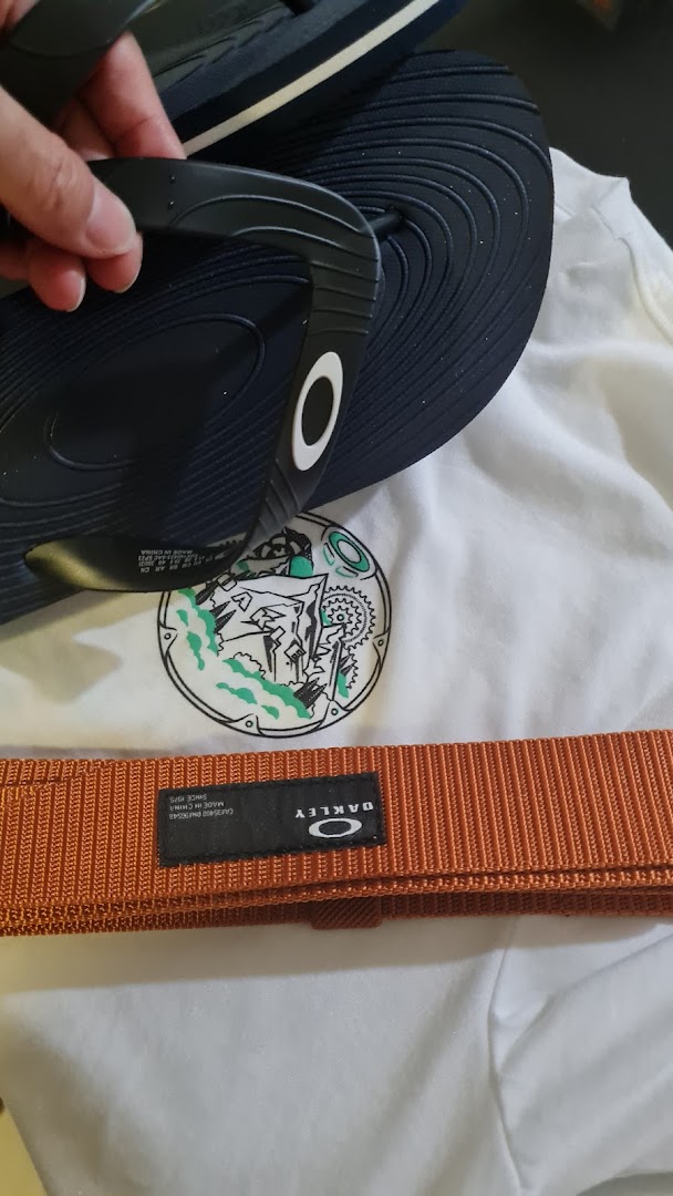 A hand holds black Oakley flip-flops, alongside an orange Oakley belt and a white graphic t-shirt.
