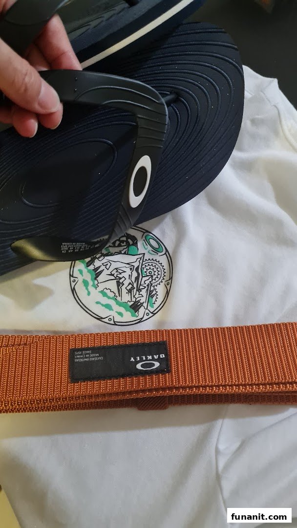 A hand holds black Oakley flip-flops, alongside an orange Oakley belt and a white graphic t-shirt.