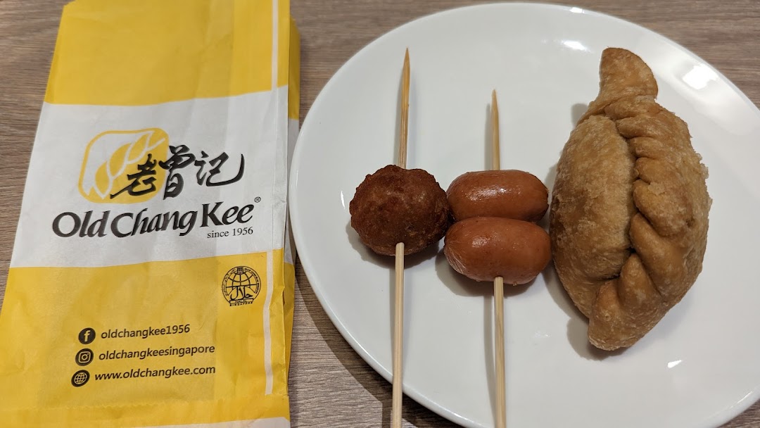 A white plate with Old Chang Kee snacks including a curry puff, fishball, and sausages, next to a branded paper bag.