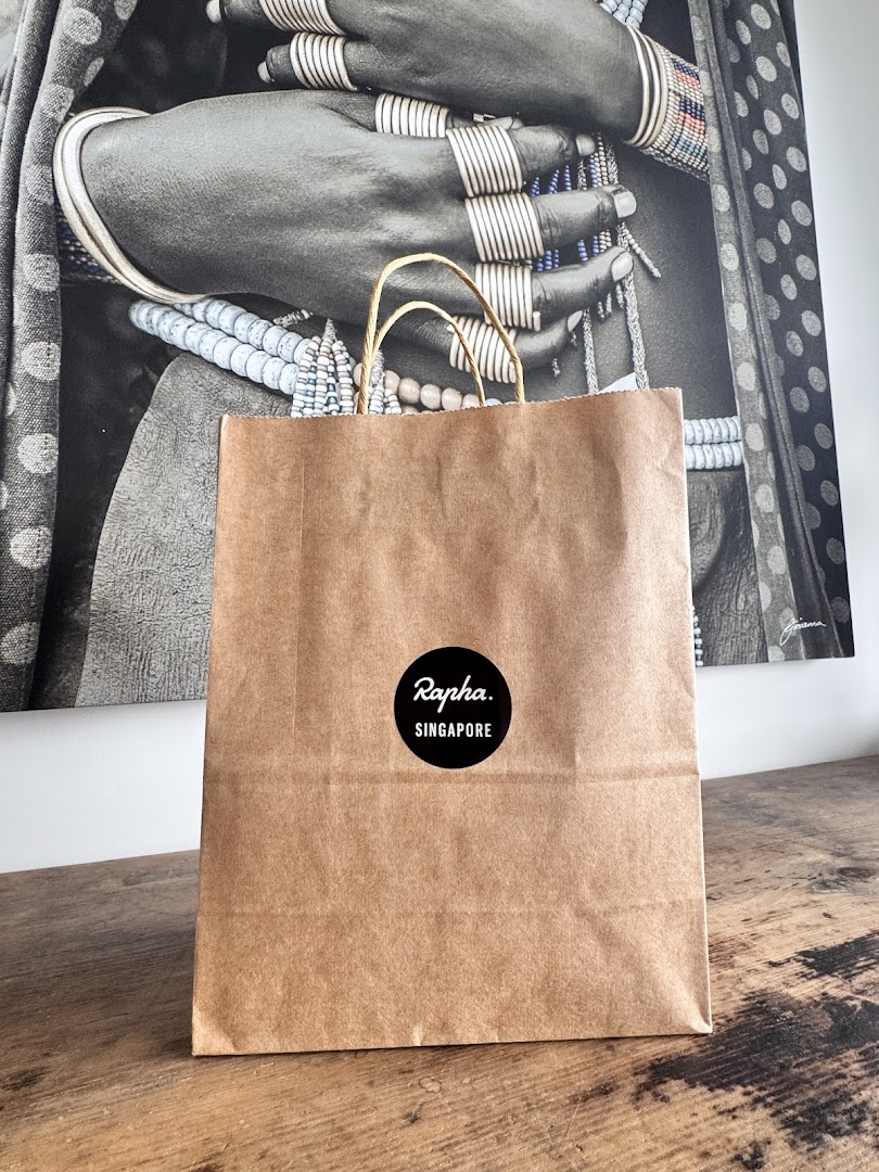 A brown paper shopping bag with a black 'Rapha. Singapore' logo sits on a wooden surface.